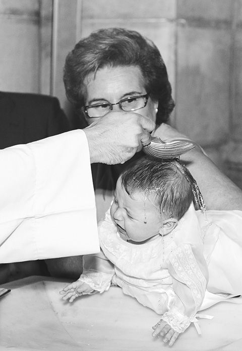 Abuela sujeta al bebé mientras recibe el bautismo, la imagen es en blanco y negro, el bebé lleva un faldón blanco y la abuela lleva gafas, la imagen fue tomada en un reportaje de bautismo.