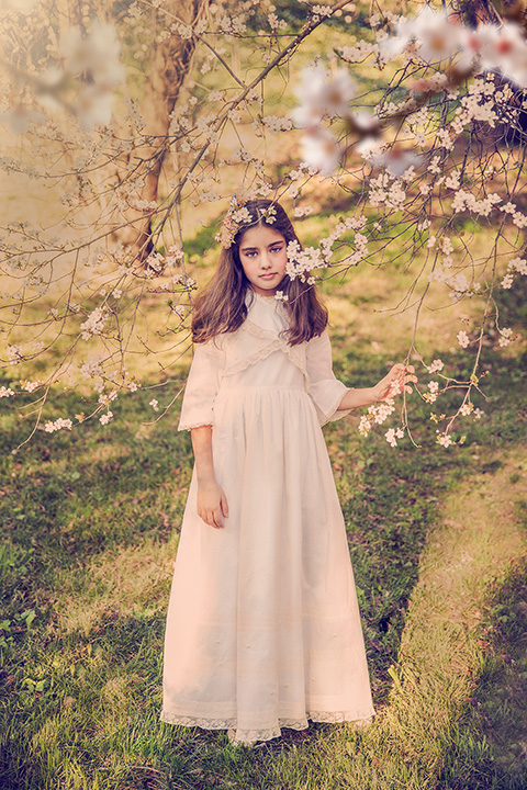Plano general de una niña con un vestido beige y tocado en la cabeza, rodeada de flores, la imagen realizada para un catálogo de comunión en exteriores.