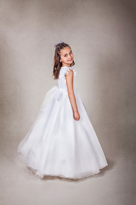 Sesiones de fotos de comunión en Madrid en estudio de fotografía de una niña con vestido blanco y un lazo detrás, tiene pelo largo que mira hacía la cámara.