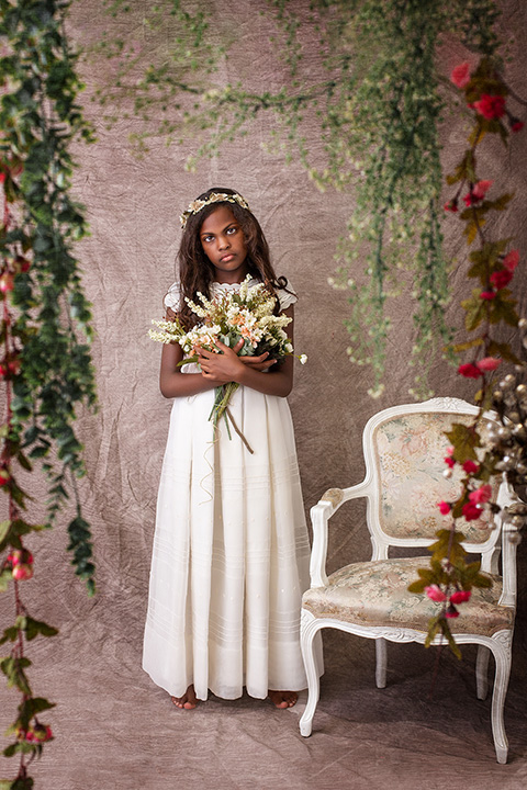Niña en su sesión de fotos de comunión en estudio, lleva un vestido blanco y un ramo entre se manos, alrededor de ella cuelgan flores rosas y a su lado tiene una silla de madera estilo clásico. 