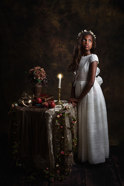 Niña morena con pelo largo, es un plano general, está de pie con un vestido blanco y una corona de flores en su cabeza, sus manos están apoyadas sobre una mesa donde hay una vela encendida, libros, manzanas y un florero. Está en un estudio de fotografía de comunión.