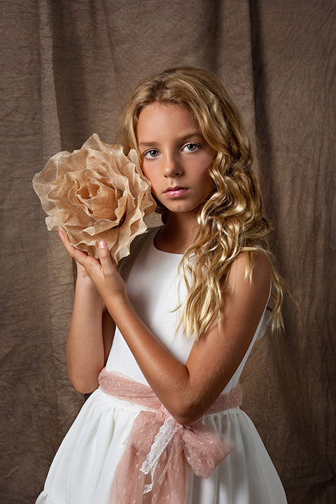 Imagen de una niña en un estudio de fotografía de Primera Comunión, lleva un vestido blanco, un cinturón rosa palo y sujeta una flor beige entre sus manos.