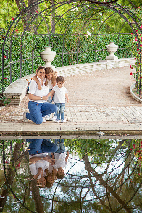Sesiones familiares en Madrid de una madre con sus hijos en el Parque del Oeste, están sobre un pequeño estanque, mirando el reflejo del agua, visten los tres camisetas blancas y vaqueros.