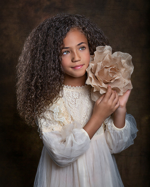 Niña con pelo rizado, vestido beige de encaje y tul, sujetando con las manos una flor de tela en una sesión de fotos infantil.
