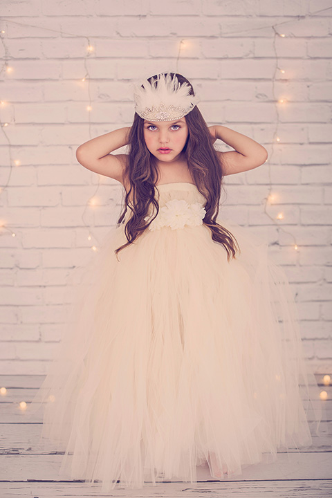 Niña de pelo castaño con un vestido largo de tul, tiene el pelo largo y lleva una cinta en la cabeza de plumas sobre un fondo de ladrillo blanco y una guirnalda de luces en un estudio de fotografía infantil en Madrid.