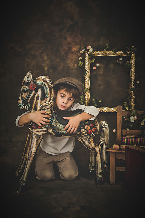 Fotografía editorial infantil de un niño sentado de rodillas, abrazando un caballo, lleva una boina y tiene una silla al lado con libros antiguos y de fondo hay un marco dorado con flores.
