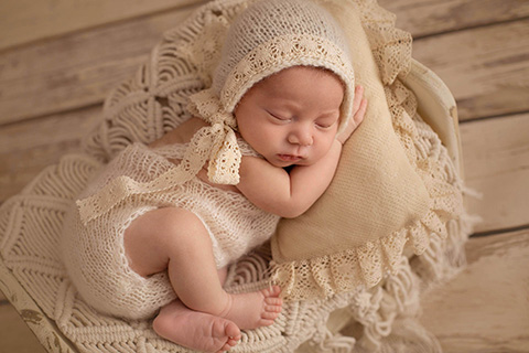 Sesión newborn de un bebé tumbado en una cama pequeña blanca, viste un body y gorro beige de encaje.