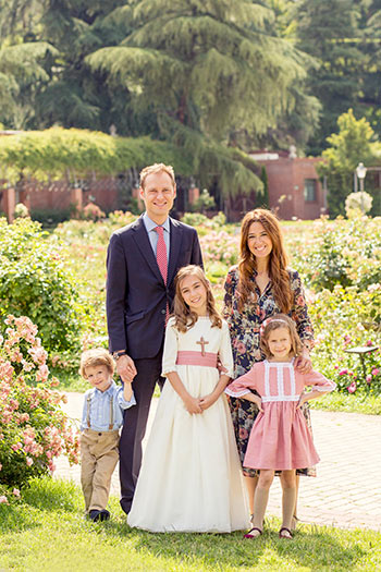 Familia al completo posan en un reportaje de comunión en Madrid, la niña mayor en su sesión de primera comunión lleva un vestido blanco largo y una cruz cuelga de su cuello. Están en un parque con flores y árboles.