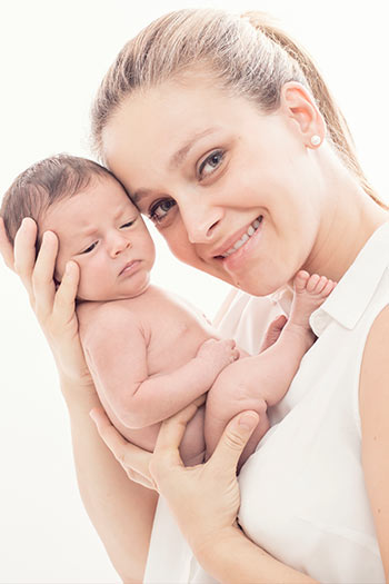 Madre cogiendo a su bebé entre sus manos y apoya su cabeza sobre él, están en su primera sesión de fotos de recién nacido en Madrid, la mamá lleva una camisa blanca y el niño está sin ropa.